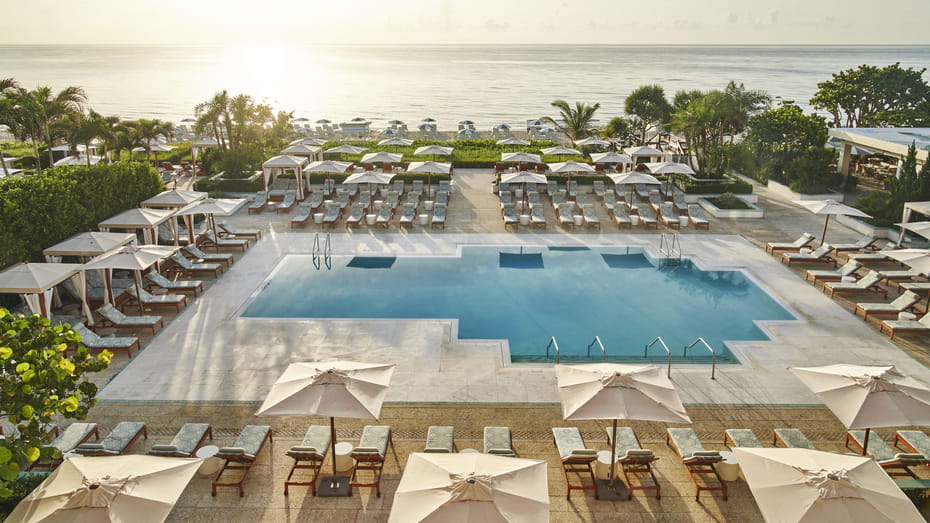 A pool surrounded by lounge chairs and large umbrellas with the ocean in the background.