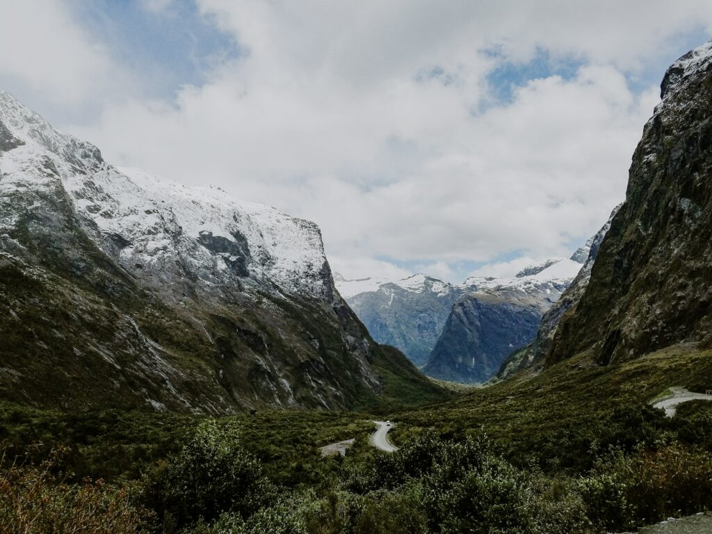 green grass field near snow covered mountain under white clouds and blue sky during daytime